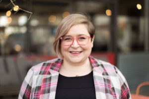 Kate has light hair and eyes. She smiles, looking at the camera. She is wearing a pink plaid coat and pink eyeglasses.