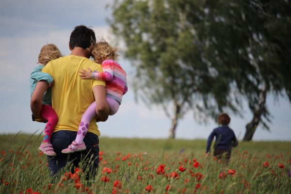A man in a yellow shirt walks through a field holding two young girls who are hugging him.